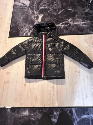 Svart Moncler puffer- Size M - Moncler svart dunjacka 🧥✨  Stilren och exklusiv svart dunjacka med blank/glansig finish 💎 Tillverkad i högkvalitativt material som både är lätt och mycket värmande 🔥 Perfekt för höst och vinter – håller dig varm samtidigt som du ser riktigt trendig ut ❄️  Jackan har en praktisk huva, smidig dragkedja med röd/vit signaturdetalj samt flera fickor med bra förvaring 🧵 Den ikoniska Moncler-loggan sitter snyggt placerad på bröstfickan och ger en lyxig känsla ⭐️  Passar perfekt till både vardag
