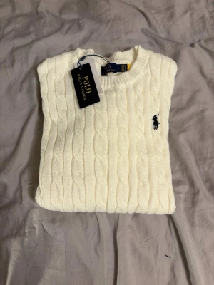 Vit kabelstickad tröja Polo Ralph Lauren - Kabelstickad vit tröja från Polo Ralph Lauren med rund halsringning och den klassiska broderade logotypen i svart på bröstet. Tröjan har långa ärmar och ribbade muddar vid ärmslut och nederkant. Perfekt för en clean och stilren look. mått: axelbredd 40cm. längd 64cm. säljer då den inte passar!
