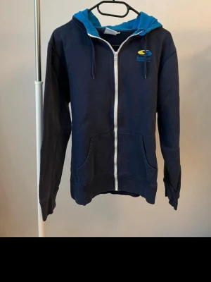 Mörkblå zip-hoodie från Svenskt kosttillskott  - Mörkblå hoodie med dragkedja från Svenskt kosttillskott. Har blå huva med dragsko, fickor fram och Svenskt kosttillskott-logga i gult och blått på bröstet samt stort tryck på ryggen. Perfekt för en avslappnad och sportig stil. Tillverkad i mjukt bomullsmaterial.
