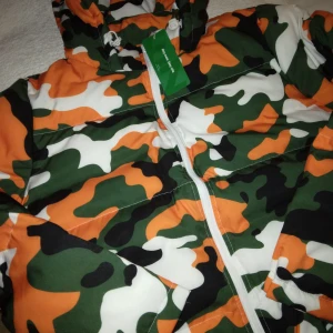 HELT NY & OANVÄND cool camouflage jacka / vinterjacka, Strlk: Medium - HELT NY & OANVÄND cool camouflage jacka / vinterjacka, Strlk: Medium/38-40. Vit/grön/orange. Vadderad och varm med fickor och luva. Fickorna stängs med lock och har tryckknapp på varje. Har pytteliten defekt framtill i form av blå liten prick från bläckpenna, syns knappt - se bilder, tex sista bilden.