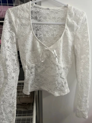 Vit spetsblus med knyt och v-ringning - Säljer en superfin vit långärmad topp i spets med v-ringning och söt knytdetalj framtill. Modellen är lätt croppad och har en romantisk känsla med transparent spetsmaterial. Perfekt att styla med jeans eller kjol för en trendig look.