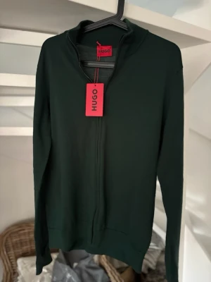 Mörkgrön ziptröja från HUGO - Snygg mörkgrön ziptröja från HUGO med hög krage och dragkedja framtill. Tröjan har en stilren design och är tillverkad i ett mjukt material som passar perfekt till lager på lager. Långa ärmar och ribbade muddar ger en clean look.
