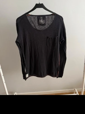 Svart långärmad tröja med ficka - Svart långärmad tröja från Xpress med rund halsringning och en liten bröstficka. Tröjan har tunna röda sömmar längs axlar och nederkant för en cool kontrast. Tillverkad i ett lätt och tunt material som känns skönt mot huden.