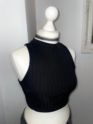 Svart ribbad croppad topp - Snygg svart croppad topp med hög hals och ribbad struktur. Toppen är ärmlös och har två knappar i nacken för stängning. Doublelined med silkigt material på insidan. Storlek XS-S. Köpt i boutique i London