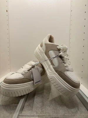 Bershka skor - Säljer nu mina helt oanvända skor ifrån bershka då dom inte kommer till användning. Skorna är vita och grå/beige mocha detaljer som ger en väldigt casual, men ändå sportig look. Dom är köpa för 32€!!