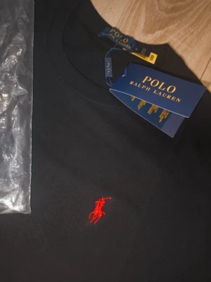 Svart t-shirt från Polo Ralph Lauren - Klassisk svart t-shirt från Polo Ralph Lauren med röd broderad logga på bröstet. T-shirten har rund hals och korta ärmar, tillverkad i mjuk bomull för en skön känsla. Perfekt basplagg med stilren design.