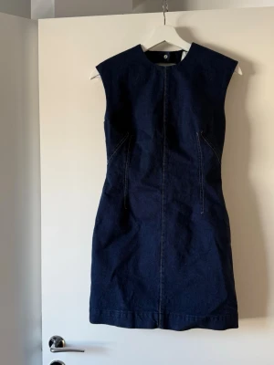 Mörkblå ärmlös jeansklänning - Snygg mörkblå jeansklänning i kort modell med markerade sömmar och öppen rygg. Klänningen är ärmlös och har en rak, något figurnära passform. Perfekt för dig som gillar en stilren och modern look.
