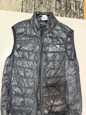 Grå dunväst från EA7 Emporio Armani - Snygg grå dunväst från EA7 Emporio Armani med hög krage och dragkedja framtill. Västen har ett stilrent quiltat mönster och logga på bröstet. Perfekt att slänga över en hoodie eller tröja för en sportig och trendig look.
