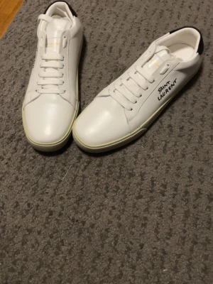 Vita Saint Laurent sneakers i läder - Snygga vita sneakers från Saint Laurent i slimmad modell med svart detalj på hälen och logga på sidan. Skorna är gjorda i slätt läder och har vita snören samt en klassisk låg siluett. Perfekta för dig som gillar stilrena och exklusiva sneakers. Pris kan diskuteras 