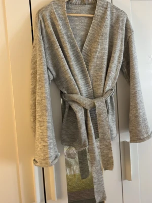 Grå stickad kofta med knytskärp - Mysig grå stickad kofta med breda ärmar och knytskärp i midjan. Koftan har öppen front och är tillverkad i mjukt material som ger en avslappnad look. Perfekt att slänga på sig över en t-shirt eller topp för extra lager. Kan användas utan knytskärp . Hittade inte storleken på den men ser ungefär ut och vara i storlek xs/s/m . Mjuk och skön använd några ggr. Pris kan diskuteras 💞