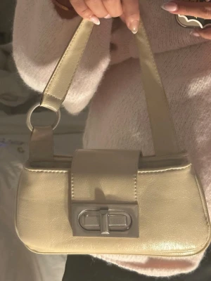 Beige axelremsväska med silverdetaljer - Trendig beige handväska i slätt konstläder med bred axelrem och silverfärgade metalldetaljer. Väskan har ett lock med vridlås framtill och dragkedja upptill för extra säkerhet. Perfekt storlek för det viktigaste och ger en stilren look.