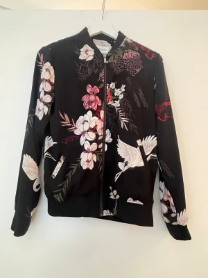 Svart jacka  med blommor & tranor - Svart bomberjacka från Gina Tricot med färgglada blommor och vita tranor tryckta över hela plagget. Jackan har dragkedja framtill, ribbade muddar och en avslappnad passform. Materialet känns mjukt och lätt, perfekt för en trendig look.