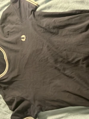 Svart Fred Perry t-shirt med vita detaljer - Klassisk svart t-shirt från Fred Perry med vita ränder på krage och ärmslut samt den ikoniska loggan broderad på bröstet. T-shirten har rund hals och är tillverkad i mjukt bomullsmaterial. Perfekt för en stilren och sportig look.