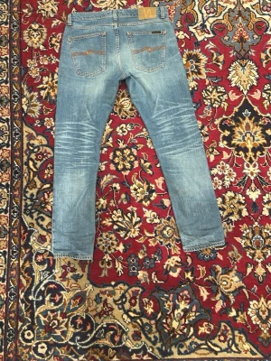 Ljusblå slim fit jeans från Nudie - Säljer ett par ljusblå slim fit jeans från Nudie Jeans med klassiska fem fickor och snygga slitningar på både fram- och baksida. Jeansen har kontrastsömmar och läderpatch bak i midjan. Perfekta för en avslappnad och trendig look.