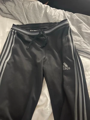 Svarta Adidas mjukisbyxor med ränder - Säljer ett par svarta mjukisbyxor från Adidas med de klassiska gråa ränderna längs benen och Adidas-logga på låret. Byxorna har dragsko i midjan och är tillverkade i ett mjukt och bekvämt material. Perfekta för chill eller träning. 