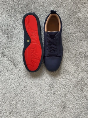 Mörkblåa Christian Louboutin skor  - Snygga mörkblå sneakers med ovandel i mocka och klassisk snörning. Insidan är beige och yttersulan är iögonfallande röd med ett mönster. Perfekta för dig som vill ha en stilren men unik look.