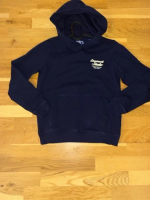 Mörkblå hoodie med tryck - Mörkblå hoodie med huva och magficka. Liten vit texttryck på bröstet. Tillverkad i mjukt material, perfekt för en avslappnad stil. Passar dig som gillar enkel och klassisk design.