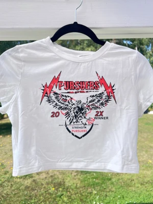 y2k croppad croppad baby tee med tryck - En y2k-vibe croppad baby tee med tryck på bröstet. Storlek L men är liten i storlek då den passar bra till mig som har S. Perfekt för dig som gillar streetstyle och grafiska prints.  (ALDRIG ANVÄND) Längd: 40 cm - Axel: 37 cm - Byst: 87 cm 