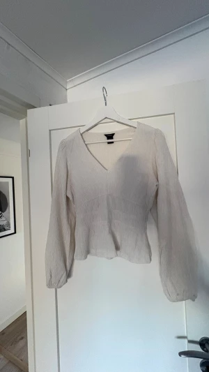 Vit v-ringad blus från Lindex - Säljer en stilren vit blus från Lindex med v-ringning och långa ärmar. Blusen har en lätt rynkad struktur och markerad midja som ger en snygg siluett. Perfekt att matcha med jeans eller kjol för en trendig look.