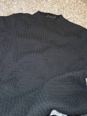 Svart stickad tröja från Dressmann - Snygg svart stickad tröja från Dressmann med diskret strukturmönster. Tröjan har rund halsringning och ribbade muddar vid halsen. Perfekt för dig som gillar stilrena och klassiska plagg i garderoben.