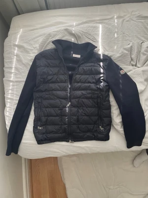 Mörkblå dunväst från Moncler - Snygg mörkblå dunväst från Moncler med quiltad front och två dragkedjefickor. Västen har stickade ärmar och hög krage för extra stilpoäng. Klassisk Moncler-logga på ärmen. Perfekt för lager-på-lager och streetwear-vibe.