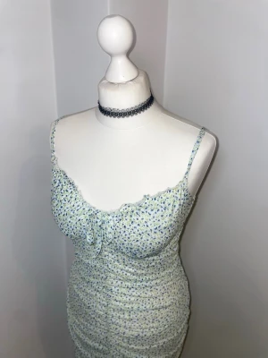 Blommig kort klänning med volang - Söt kort klänning med smala axelband och blommigt mönster i blått och grönt på vit botten. Klänningen har volangkant vid bysten och en liten knytning framtill. Figurnära passform och rynkade detaljer längs sidorna ger en feminin siluett. Perfekt för sommardagar.