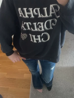 Svart Alpha Delta Chi hoodie - Cool svart hoodie med vit text 'Alpha Delta Chi' och ett leopardmönstrat hjärta på bröstet. Oversized passform och ribbade muddar vid ärmslut och nederkant. Perfekt för dig som gillar college-vibe och streetstyle.