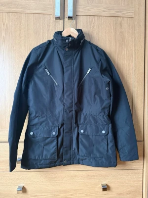 Svart parkasjacka med huva - Snygg svart parkasjacka med hög krage, justerbar huva och dragkedja framtill. Två bröstfickor med dragkedja och två stora fickor med lock och knapp framtill. Jackan har långa ärmar och en rak passform, perfekt för kyliga dagar.