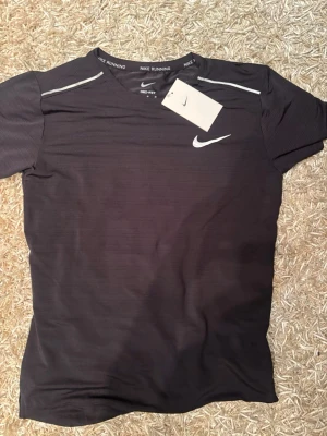 Svart Nike Dri-FIT löpartröja - Säljer en svart löpartröja från Nike med Dri-FIT-material som håller dig torr under träningen. Tröjan har vita detaljer på axlarna och ett vitt Nike Swoosh på bröstet. Perfekt för löpning och annan träning. Rund hals och korta ärmar.