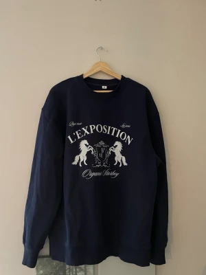 Mörkblå hoodie L'EXPOSITION - Snygg mörkblå hoodie från Organic Starboy med tryck framtill där det står L'EXPOSITION och motiv av två hästar och ett lejon. Klassisk passform med långa ärmar och rund hals. Perfekt för dig som gillar streetwear och vill sticka ut.