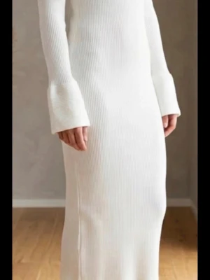 Vit ribbad långklänning - Säljer en vit ribbad långklänning med långärmad design och utsvängda ärmslut. Klänningen har en kroppsnära passform och är tillverkad i ett mjukt stickat material som känns både trendigt och bekvämt.