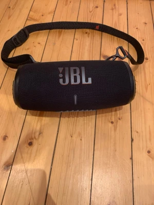 JBL svart bärbar högtalare - Snygg svart bärbar högtalare från JBL med robust tygmaterial och cylinderformad design. Utrustad med axelrem för enkel transport och tydliga knappar för volym, Bluetooth och ström. Perfekt för att ta med sig musiken överallt. Högtalaren är vattentät och går att använda var som helst. Hör av dig vid minsta fråga eller för att diskutera priset.