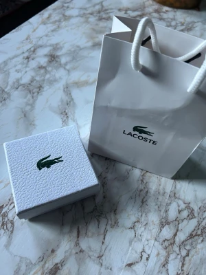 Lacoste halsband i silverfärg - Stilrent halsband från Lacoste i silverfärg med tunn kedja och klassisk krokodilberlock. Kommer i original ask och tygpåse med Lacoste-logga. Perfekt för dig som gillar minimalistiska accessoarer och vill ha något från ett ikoniskt märke.