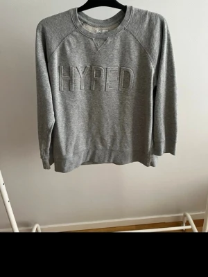 Grå sweatshirt med HYPED-tryck - Grå sweatshirt i mjukt bomullsmaterial med texten 'HYPED' broderat på bröstet. Klassisk rund halsringning och långa ärmar med ribbade muddar. Perfekt för en avslappnad och trendig look.