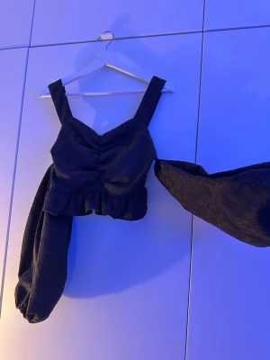 Svart topp med puffärmar och volang - Trendig svart topp med breda axelband, volang nedtill och stora puffärmar. Toppen har en lätt rynkad detalj framtill och är perfekt för dig som vill sticka ut. Materialet känns lite tjockare och har en diskret struktur. Snygg att styla med både jeans och kjol.