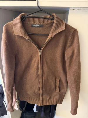 Brun ziptröja från Dressmann - Brun stickad ziptröja från Dressmann med ribbad krage och muddar. Tröjan har två fickor framtill och dragkedja hela vägen. Perfekt för lager på lager och enkel att matcha med jeans eller chinos.