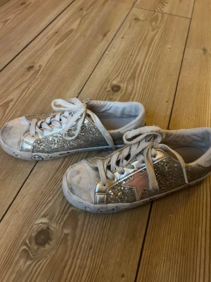 Glittriga silvriga sneakers med stjärna - Säljer ett par sneakers med silverglittriga sidor, vita snören och en rosa stjärna på sidan. Skorna har coola handritade symboler på sulan och en klassisk låg profil. Perfekta för dig som vill sticka ut lite extra!