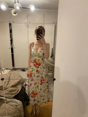 Blommig sommarklänning med smala band - Söt långklänning med smala axelband och blommigt mönster i rött, gult och grönt på vit botten. Klänningen har en lätt v-ringad urringning och en luftig passform, perfekt för varma dagar. Materialet känns som bomull eller linne.