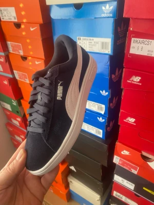 Svarta Puma sneakers med vit detalj - Snygga svarta sneakers från Puma med klassisk vit logo och vit sula. Skorna har snörning och är tillverkade i mocka med syntetdetaljer. Perfekta för dig som vill ha en stilren och sportig look.