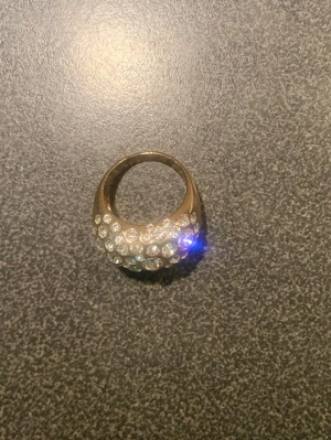 Guldfärgad ring med glittrande stenar - Snygg guldfärgad ring med massor av glittrande, klara stenar på ovansidan. Ringen har en rundad, chunky form som verkligen sticker ut och fångar ljuset. Perfekt för dig som gillar att addera lite extra bling till din stil. Strl 19 efter mätning 