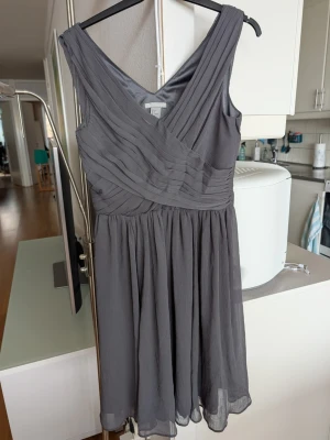 Grå omlottklänning från H&M - Snygg grå klänning från H&M med omlottdesign och v-ringning. Klänningen har breda axelband, veckade detaljer upptill och ett lätt, flygigt tyg i kjolen. Perfekt för dig som gillar stilrena och eleganta plagg. Dragkedja i sidan.