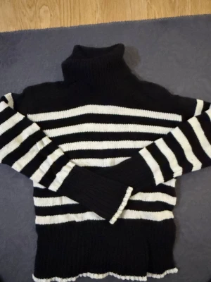 Svartvit randig polotröja stickad - Stickad polotröja i svart och vitt med breda horisontella ränder. Tröjan har hög polokrage, långa ärmar och ribbade muddar samt nederkant. Perfekt för en chill och stilren look.