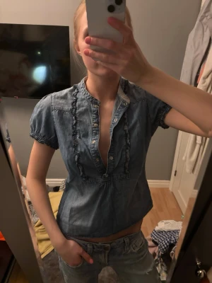 Blå jeansblus med volangdetaljer - Säljer en blå jeansblus med korta puffärmar och volangdetaljer längs knäppningen. Bär S 