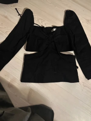 Svart croppad topp med snörning - Snygg svart croppad topp med långa puffärmar och trendig snörning framtill. Toppen har en öppen detalj vid midjan som ger en edgy look. Perfekt för dig som vill sticka ut lite extra.