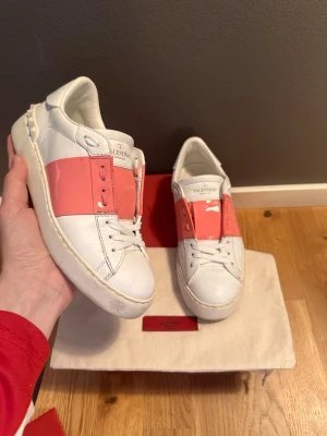 Valentino sneakers med rosa detalj - Snygga vita sneakers från Valentino med en bred rosa panel över snörningen. Skorna har klassisk låg profil, rund tå och vita snören. Tillverkade i skinn för en lyxig känsla och har subtila logodetaljer på plösen. Perfekta för dig som vill sticka ut med stil.
