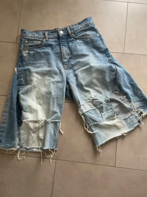Ljusblåa slitna jeansshorts som är hemgjorda! - Unika ljusblå jeansshorts med råa, fransiga kanter och stora slitningar framtill. De har en loose passform och klassisk femficksdesign. Perfekta för dig som gillar en edgy och avslappnad stil.