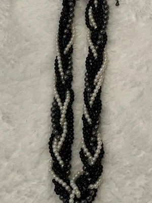 Flätat halsband med svarta och vita pärlor - Snyggt flätat halsband med flera rader av svarta, vita och grå pärlor. Halsbandet har en tvinnad design som ger en cool kontrast mellan färgerna. Perfekt accessoar för att lyfta din outfit med en unik touch. Materialet är troligen plast eller akryl.