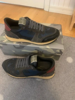 Valentino Garavani sneakers blå/svart - Snygga sneakers från Valentino Garavani i blått och svart med detaljer i vinrött och grått. Skorna har en beige sula och är tillverkade i en mix av mocka och textil. Klassisk låg modell med kamouflagemönster och grå skosnören.