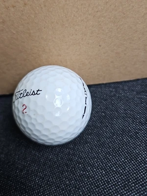 Vit Titleist golfboll - En vit golfboll från Titleist med texten 'Titleist' i svart och ett rött frågetecken tryckt på sidan. Bollen har klassisk dimple-yta och märkningen 'NXT Tour' syns på kanten.