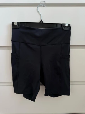 Svarta träningstights-shorts från Röhnisch - Säljer ett par svarta högmidjade träningstights-shorts från Röhnisch. De har praktiska sidofickor och är tillverkade i ett stretchigt syntetmaterial som passar perfekt för träning. Klassisk design med diskret logga framtill.